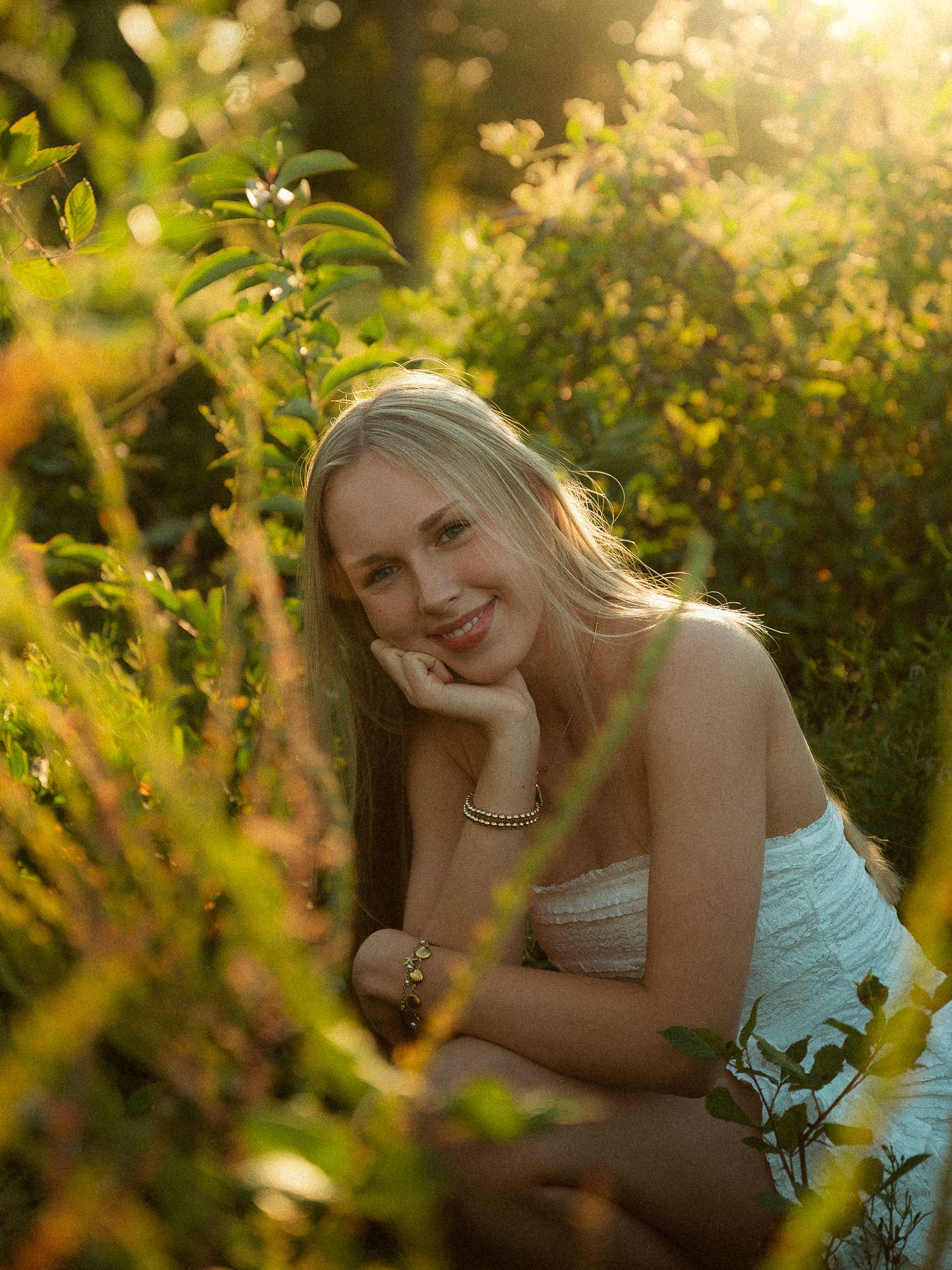 Young blonde woman smiling in golden hour sunlight surrounded by lush green garden foliage, wearing a white top and gold bracelet.