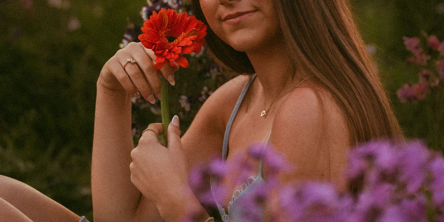 Young woman sitting in a flower garden holding a red gerbera daisy, surrounded by colorful blooms.