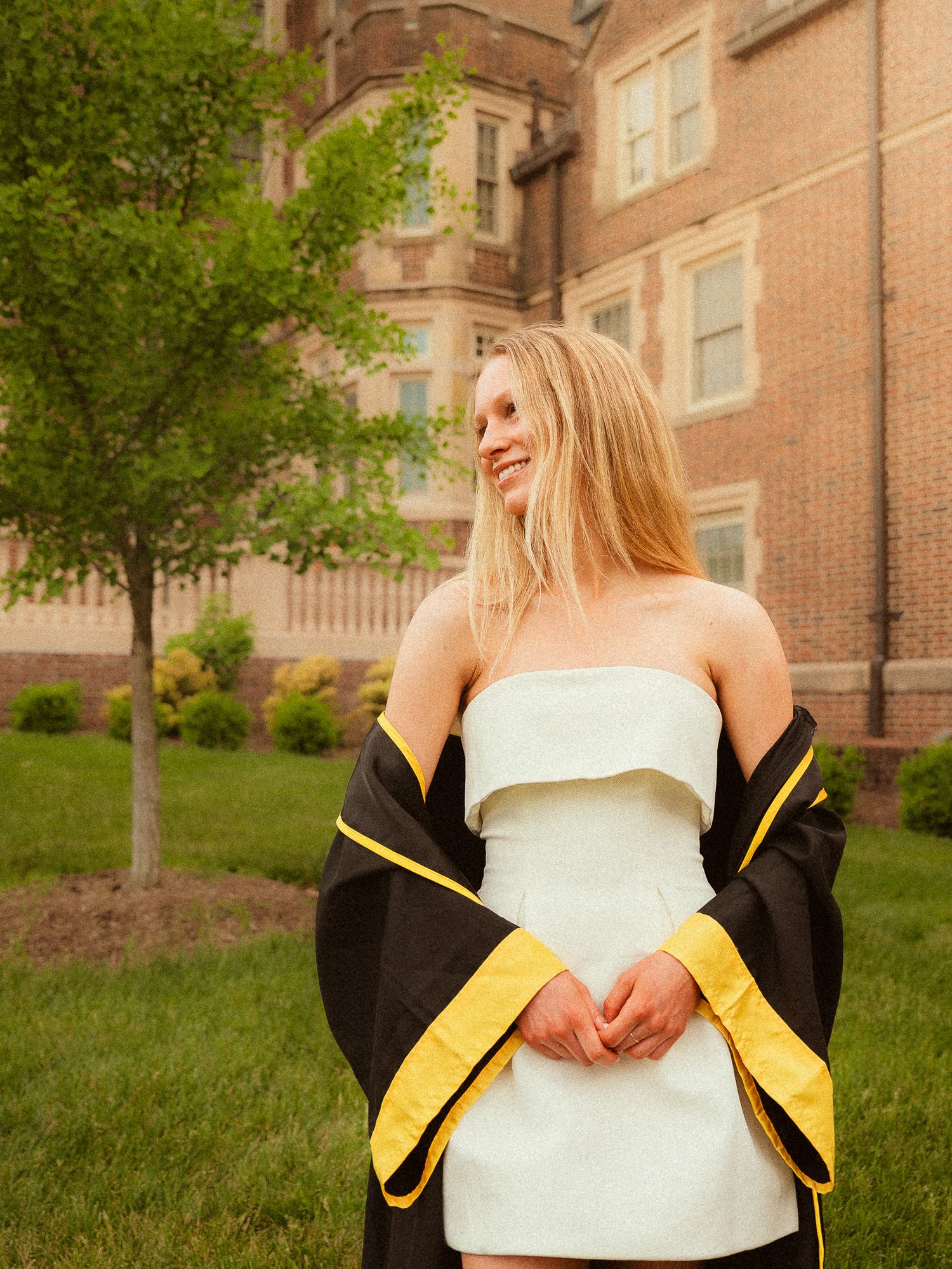 A smiling blonde woman in a white strapless dress holds a black and gold graduation gown outside a brick university building.