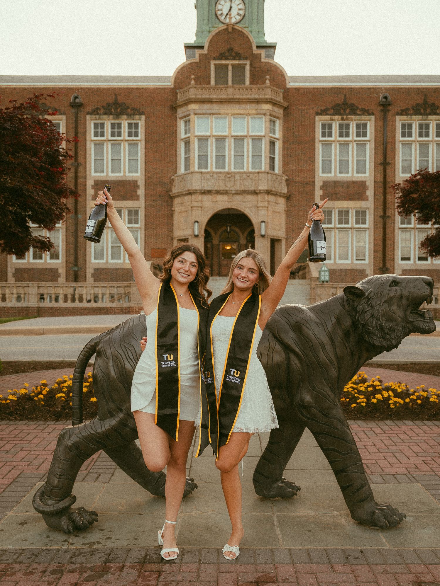 Two college graduates in caps and gowns celebrate with raised bottles in front of a brick university building with a bear statue.