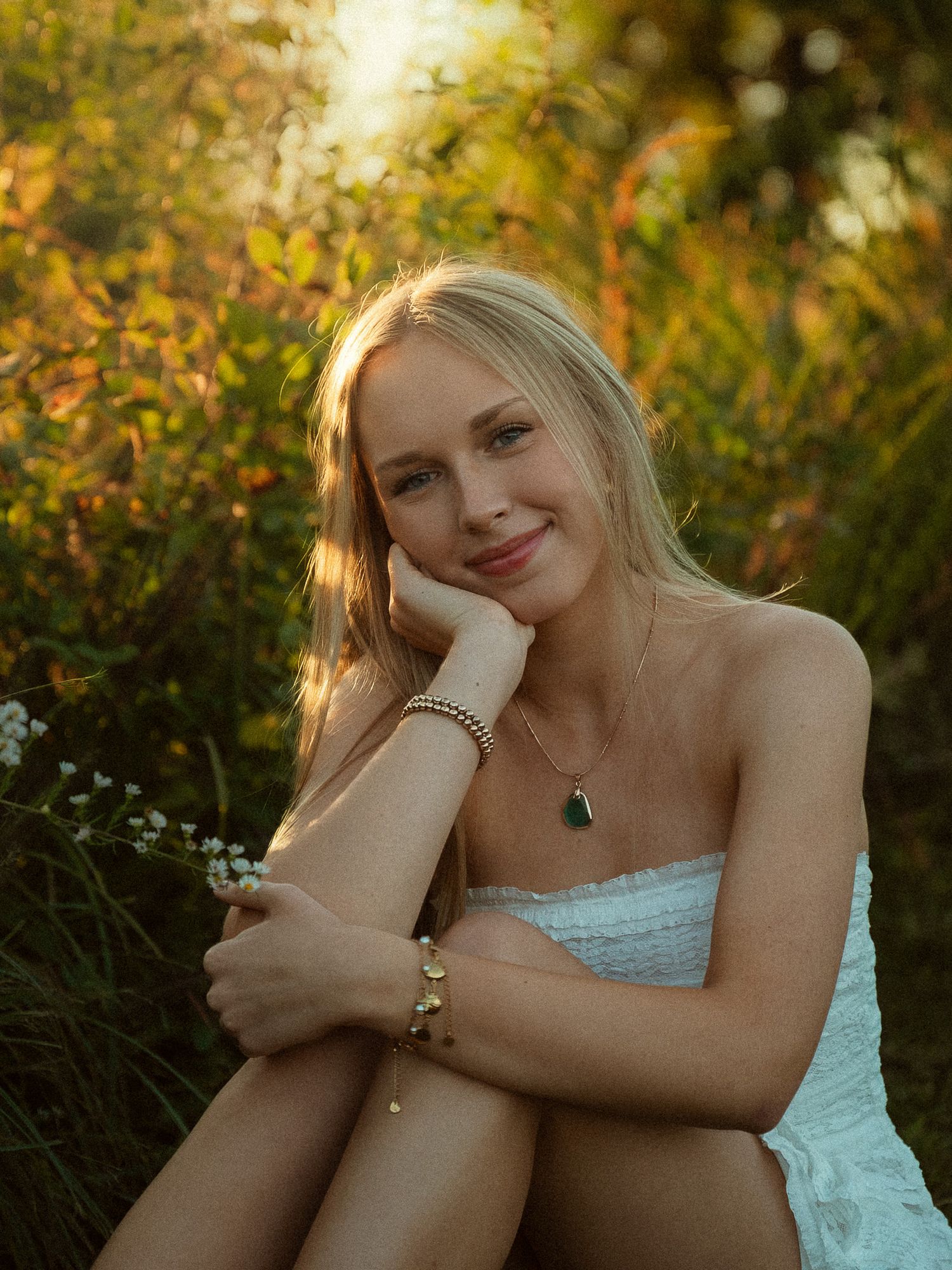 Blonde woman in white dress sitting outdoors in golden sunlight, wearing a green pendant necklace and gold bracelet.