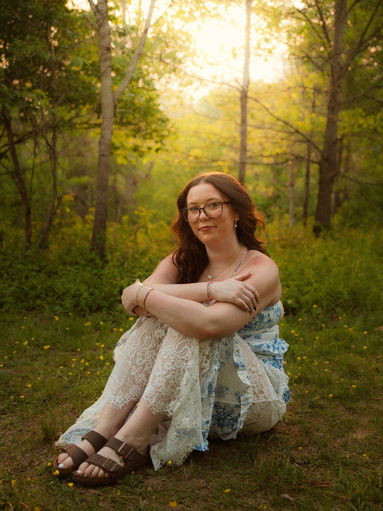 Young woman in lace dress sitting on grass in golden sunlit forest clearing, wearing glasses and sandals.