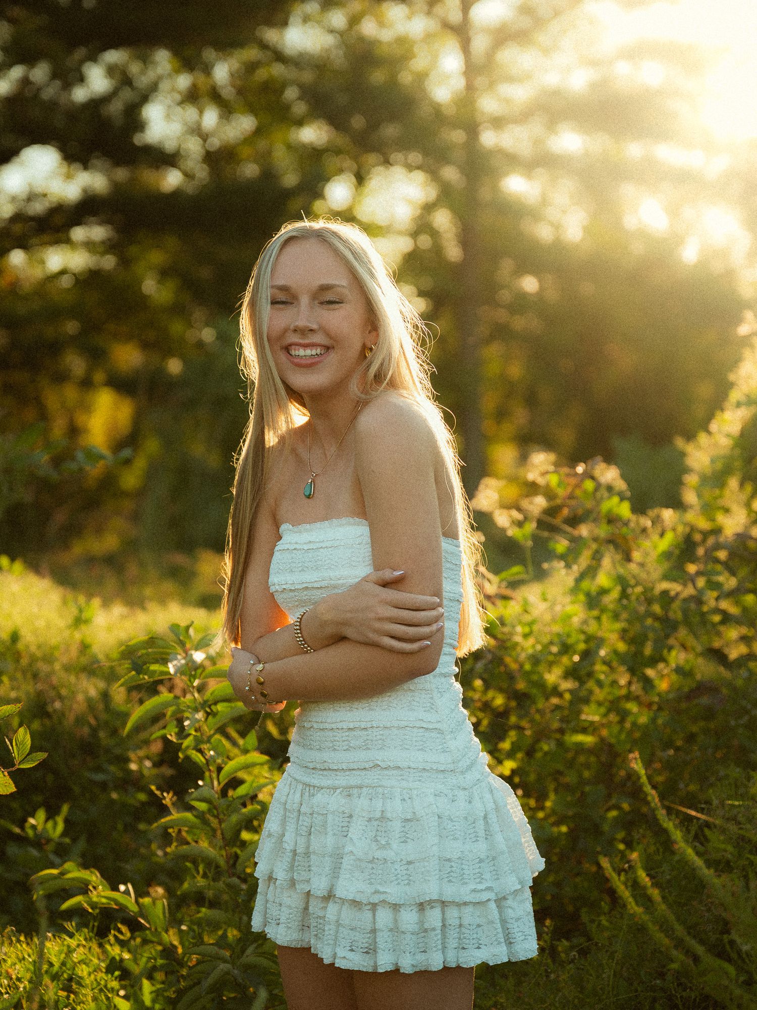 Young blonde woman smiling in white ruffle mini dress, golden sunset backlight, lush green garden background.