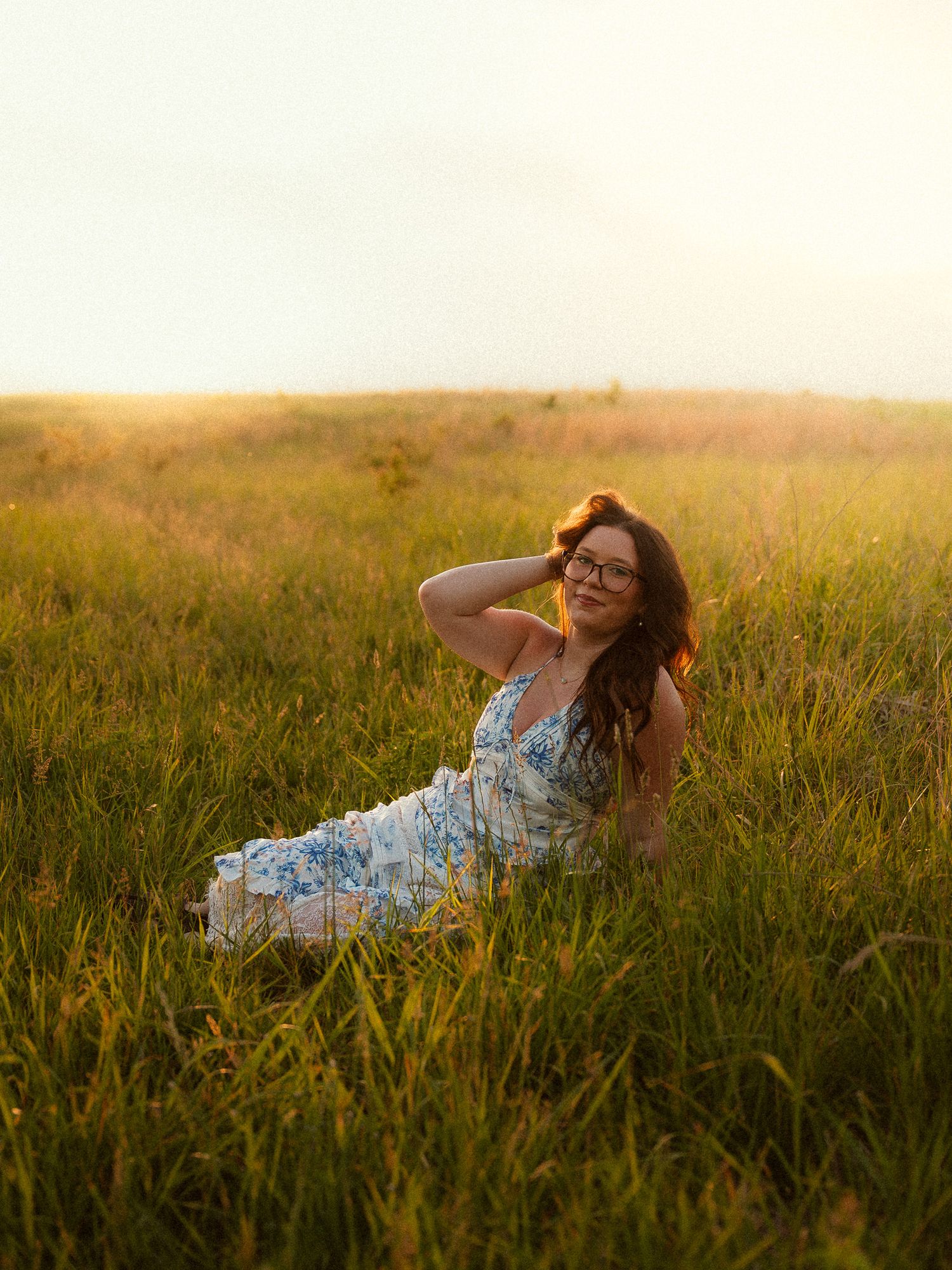 Woman in floral dress sitting in golden sunlit meadow, posing with hand in hair during sunset.