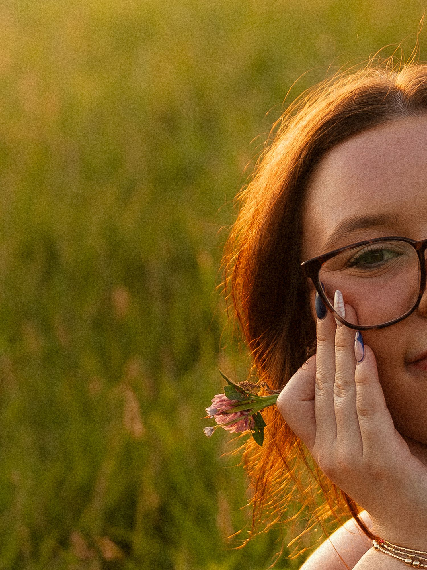 Young woman with glasses holding a pink wildflower near her face in a sunlit green field.