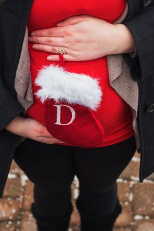Hands resting on a red Christmas stocking with white trim showing an wedding ring over pregnant woman