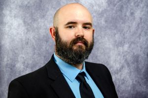 Professional studio portrait with dark beard wearing suit and blue tie.