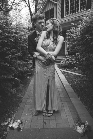 A black and white photograph of two people embracing in formal attire on a brick pathway beside a house.