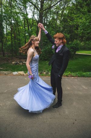 Two dancers in formal attire twirl together on a paved area surrounded by lush green trees and foliage.
