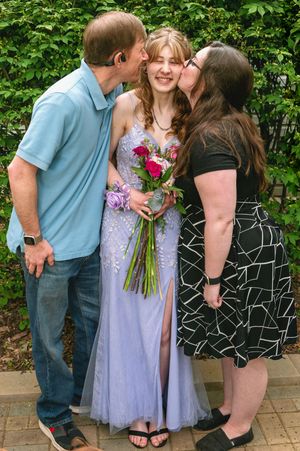 Parents kiss their daughter in a lavender prom dress as she holds a colorful bouquet of flowers against green foliage.