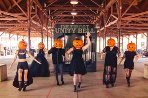 Five figures in vintage style dresses posing under a Unity Bell sign wearing pumpkin heads