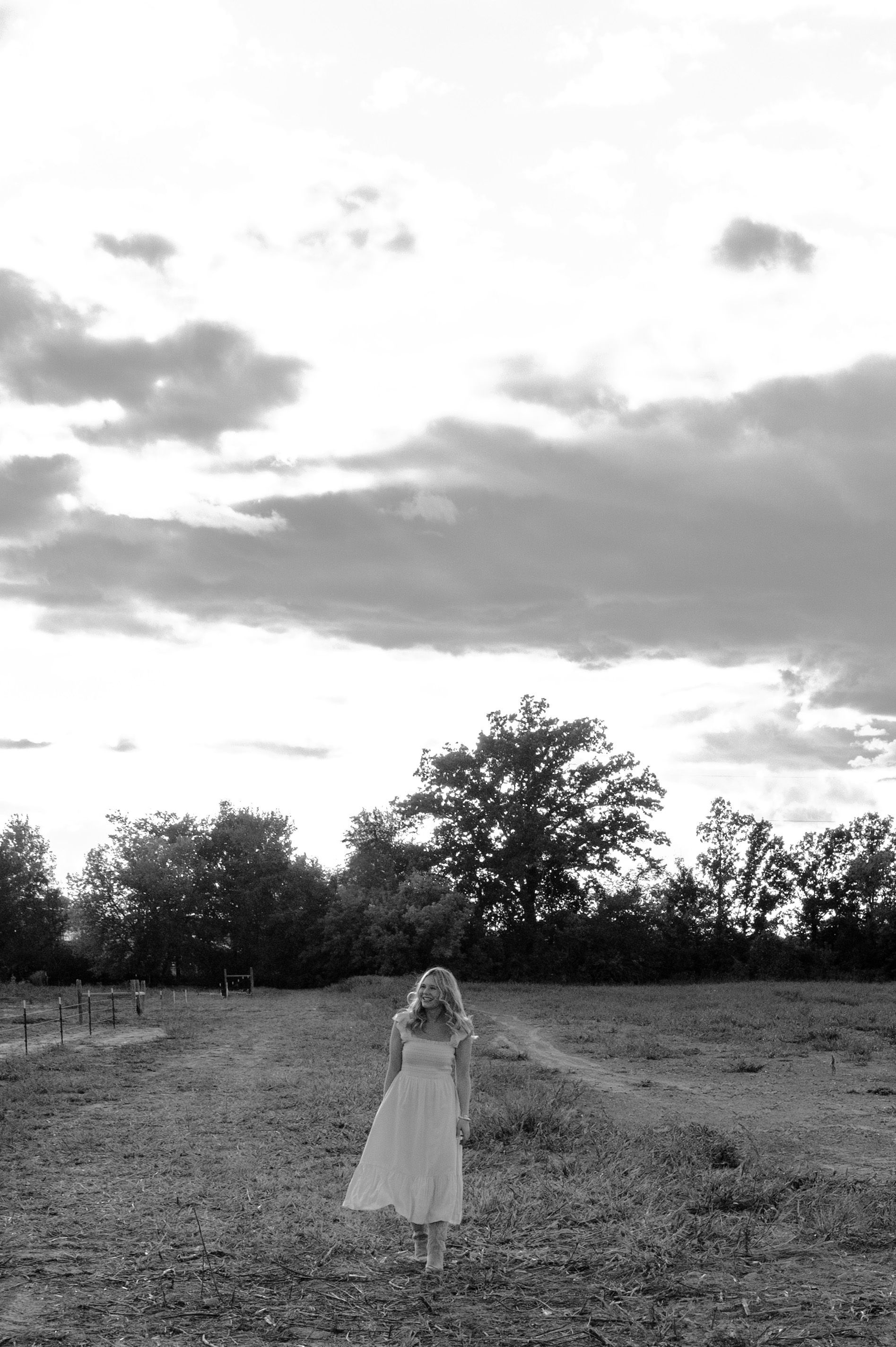 A person in a flowing white dress stands in a field at sunset while dramatic clouds drift overhead.
