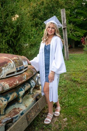 A graduation photo session featuring a white cap and gown with a rustic vintage car backdrop in a garden setting.