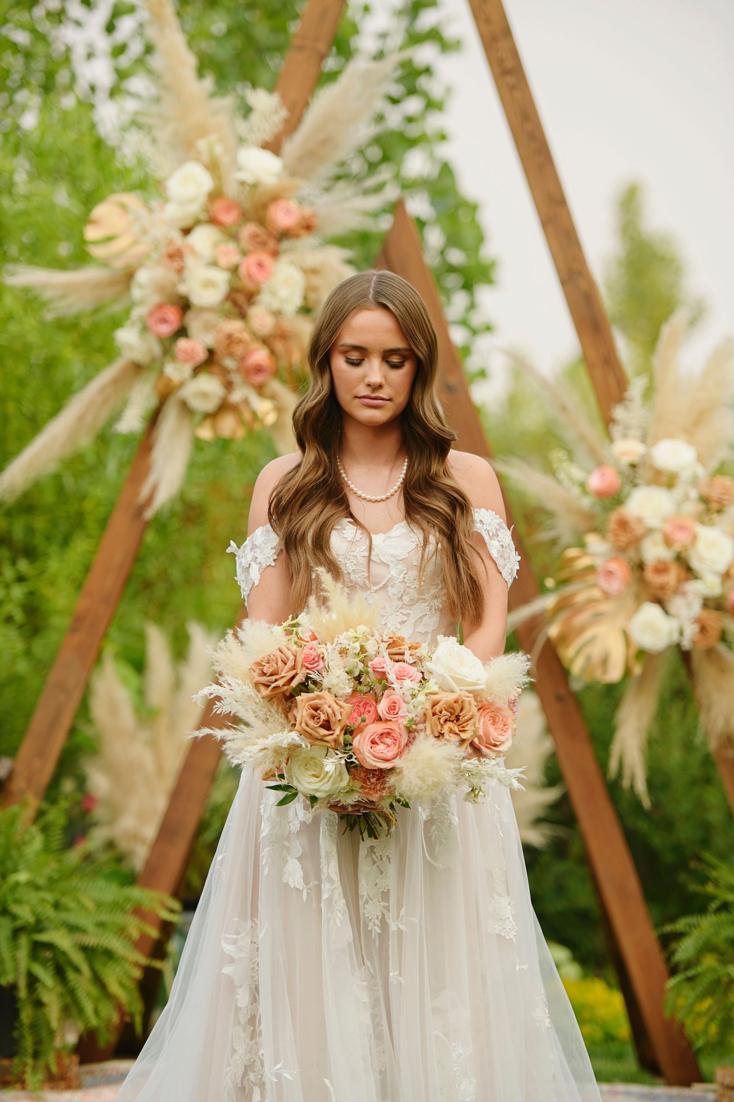 Beautiful outdoor wedding ceremony setup with wooden triangle arch decorated with pampas grass and peach-colored floral arrangements.