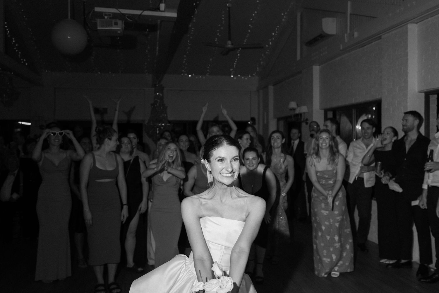 A bride in a white gown smiles while surrounded by guests dancing at a wedding reception.