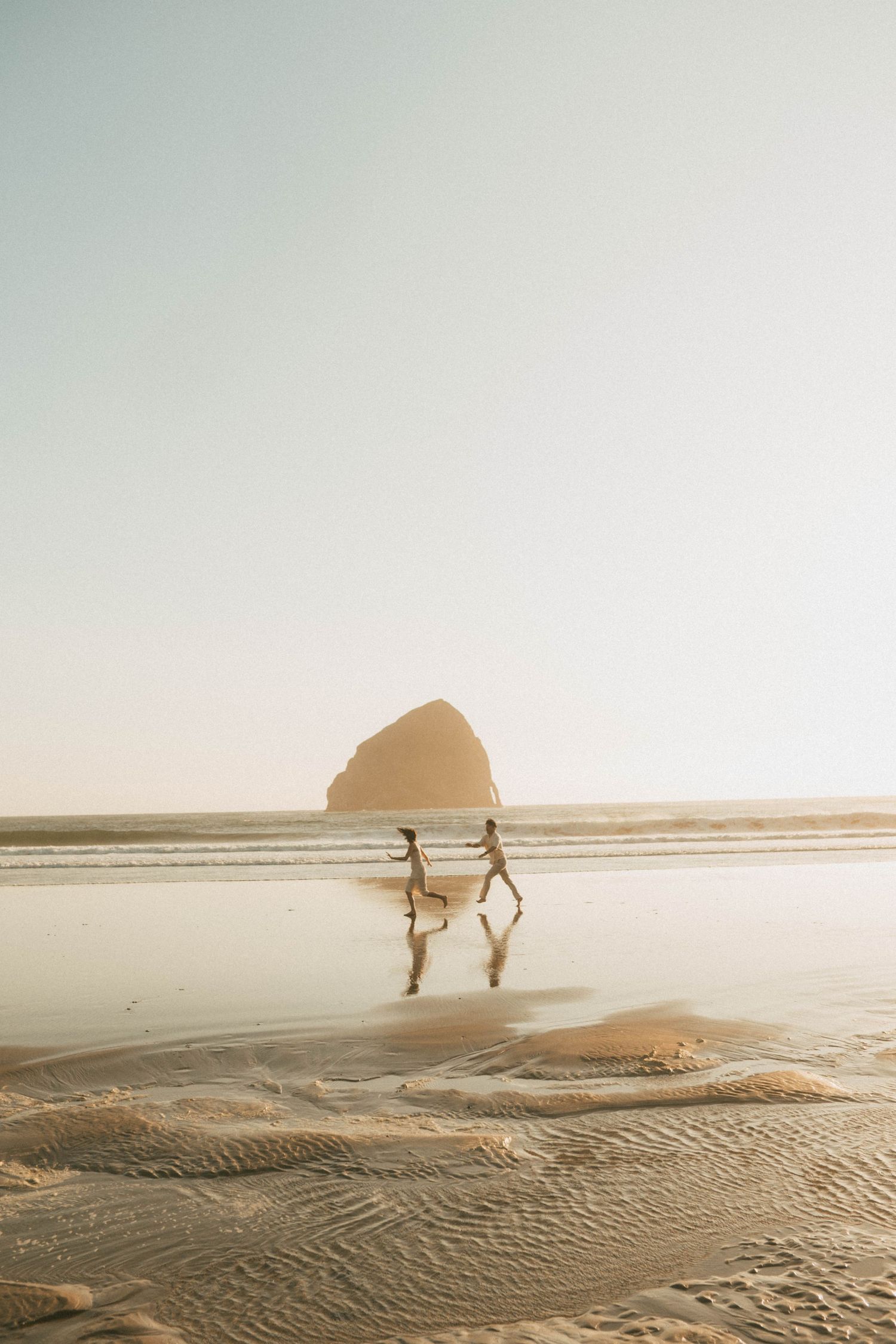 Two people share a tender moment sitting together on coastal rocks at sunset in muted golden light.