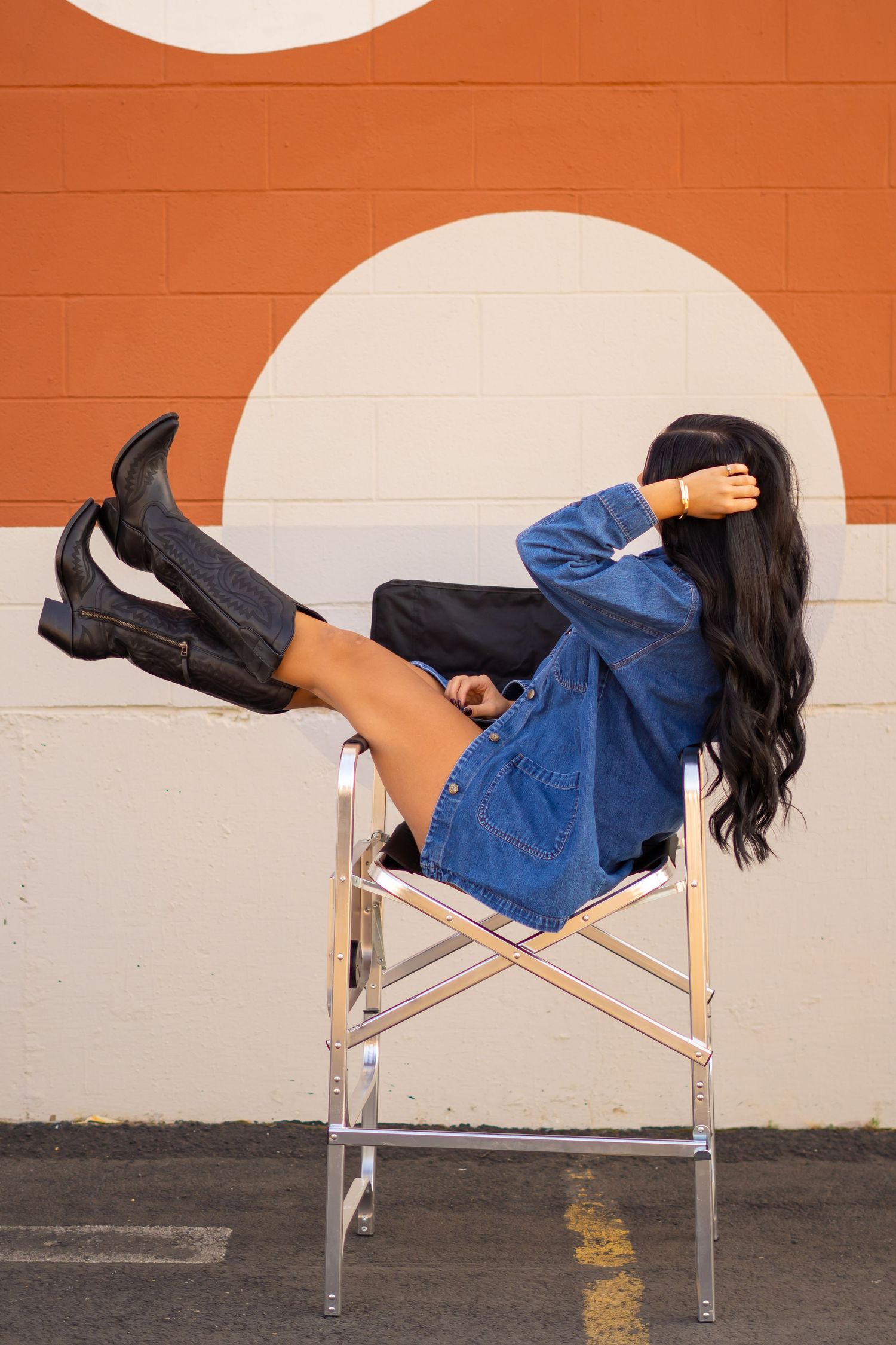 Stylish woman in denim and boots posing playfully on director's chair against orange and white wall.