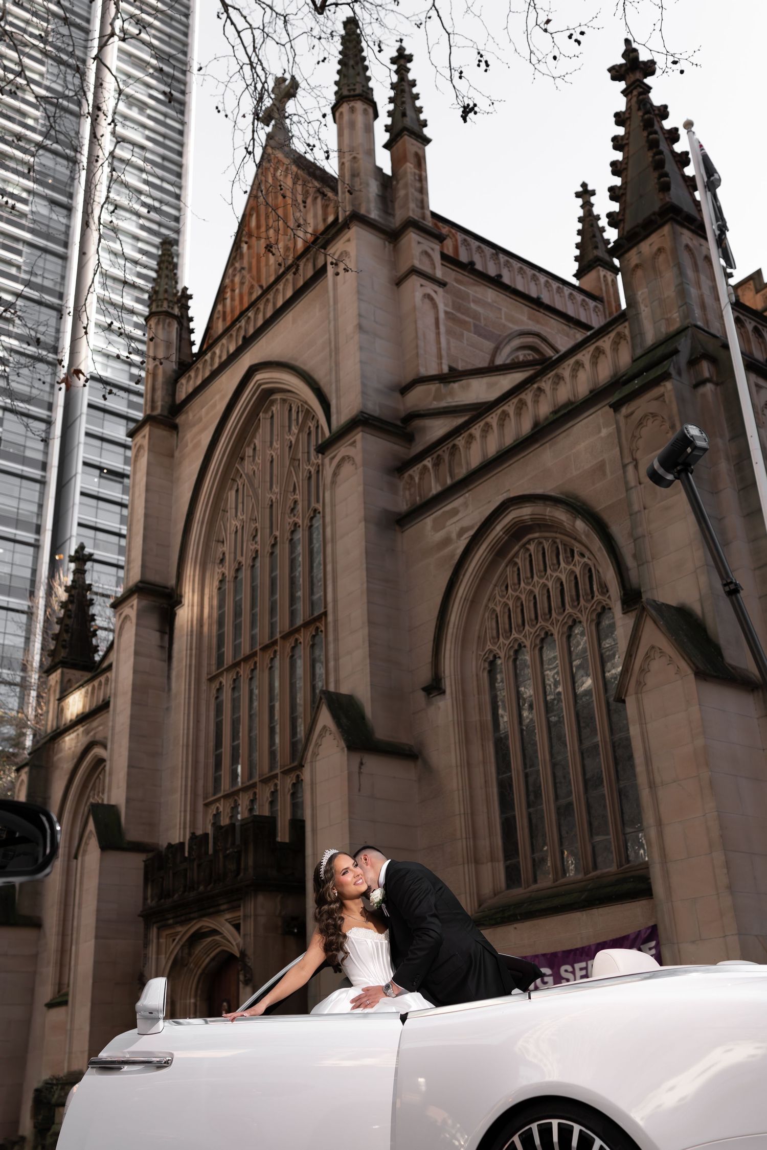 Bride and groom kissing beside white convertible car outside Gothic sandstone church.