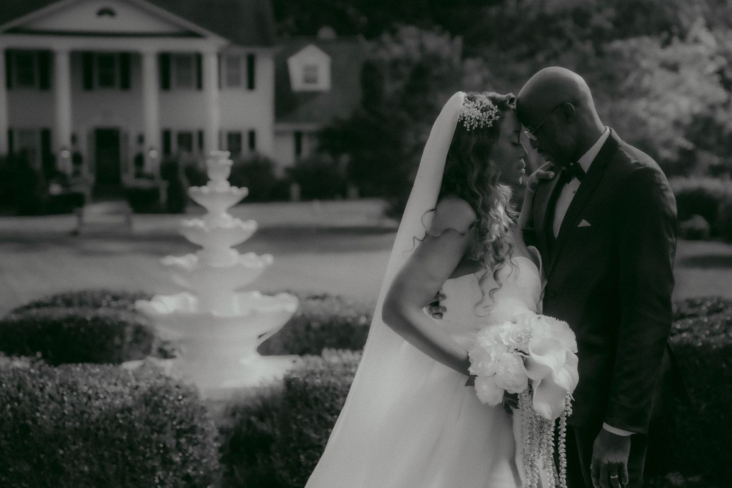 Black and white wedding photo showing romantic moment between a bride and groom in front of a stately mansion and fountain.