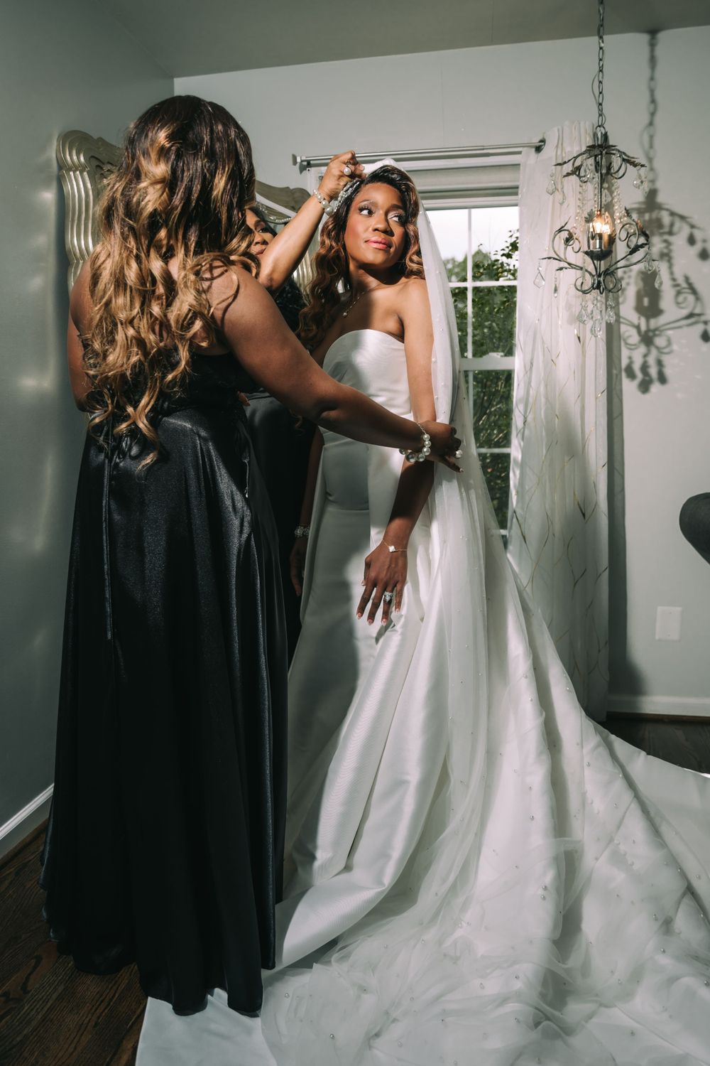 Bridesmaid in black dress helps adjust bride's elegant white wedding gown in sunlit dressing room with chandelier.