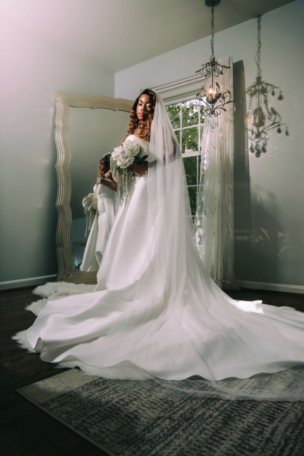 A stunning wedding gown with a long flowing train elegantly displayed in a bright room with a chandelier and window.