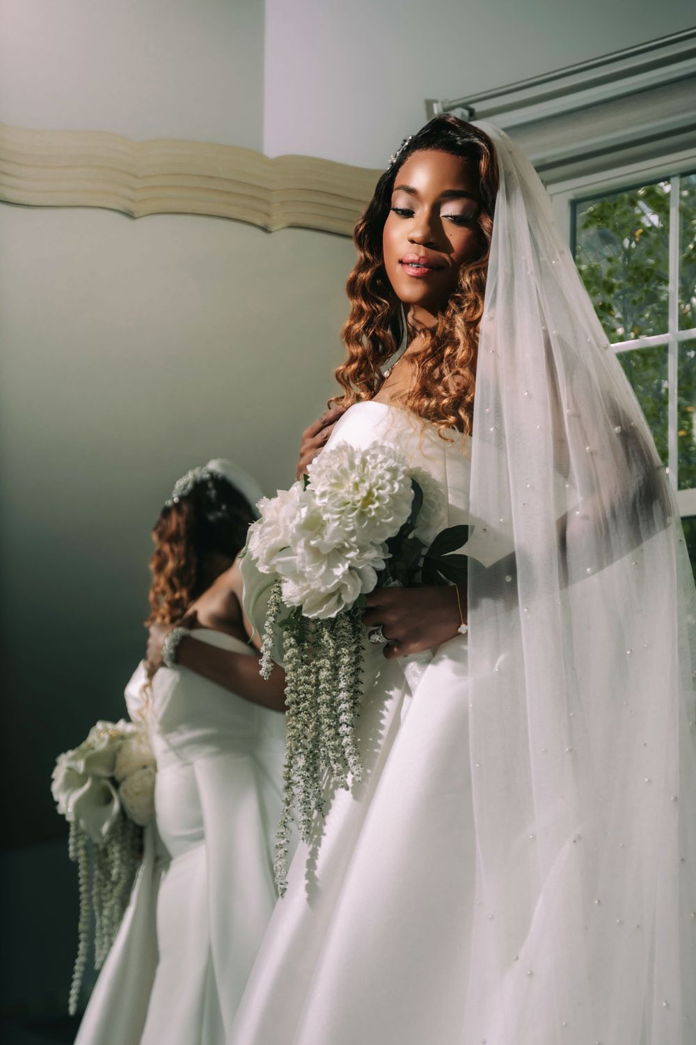 A bride in an elegant white wedding gown holds a cascading bouquet while standing near a mirror in natural light.