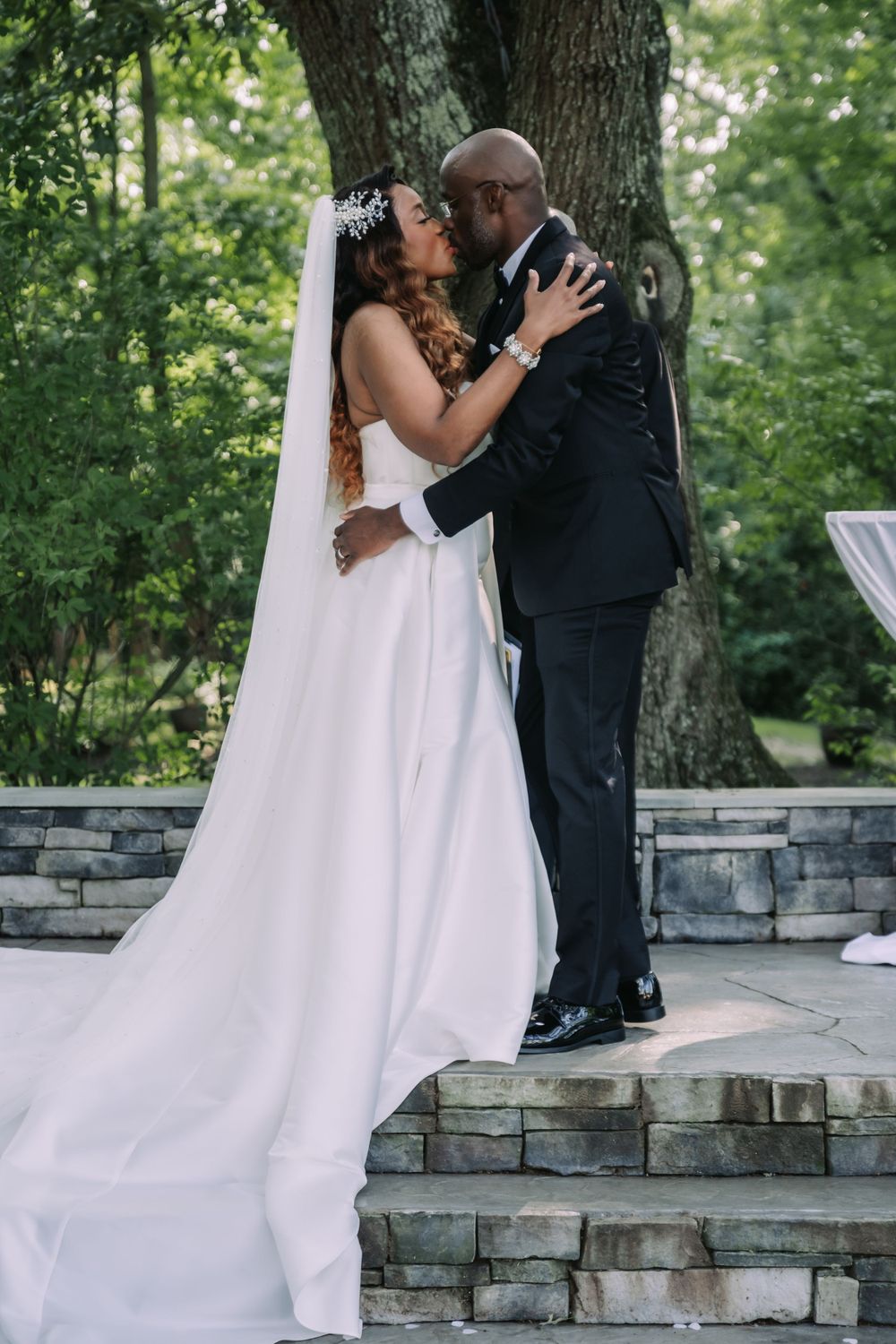 A newlywed couple shares a romantic moment on stone steps in a lush outdoor garden setting with flowing white wedding dress.