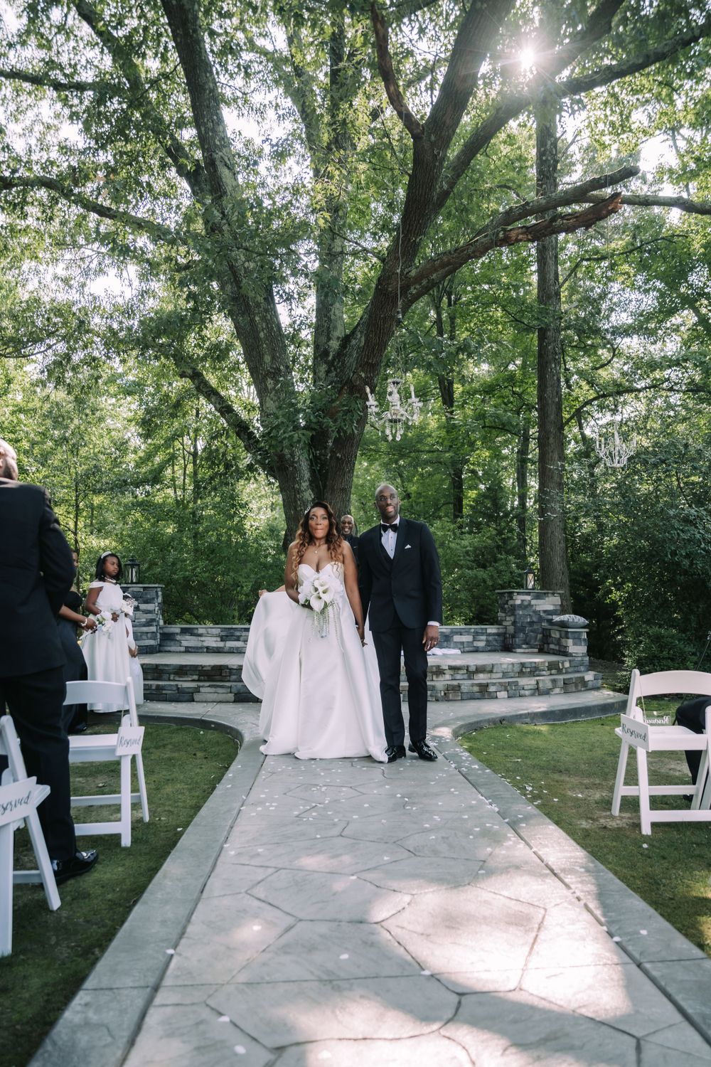 A couple stands on a stone pathway during an outdoor wedding ceremony surrounded by tall trees and white chairs.
