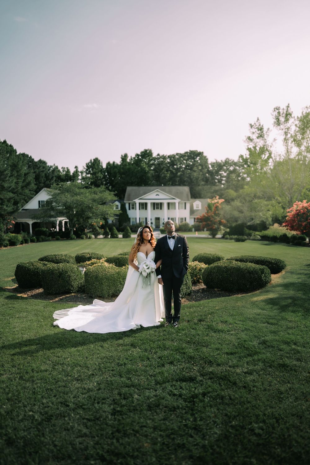 A couple in wedding attire stands on a manicured lawn with a grand white southern-style mansion in the background.