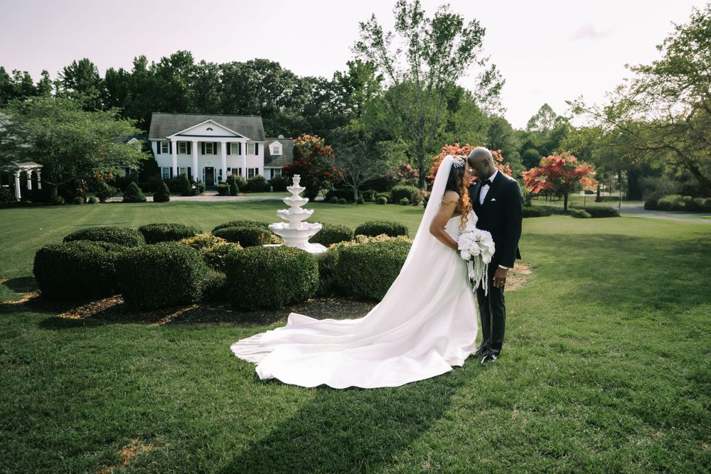 A couple shares an intimate moment on a manicured lawn with a grand southern mansion and blooming trees in the background.