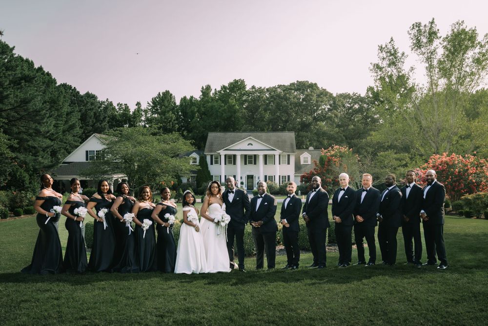 A wedding party in black and white formalwear poses on a manicured lawn in front of a classic Southern mansion.