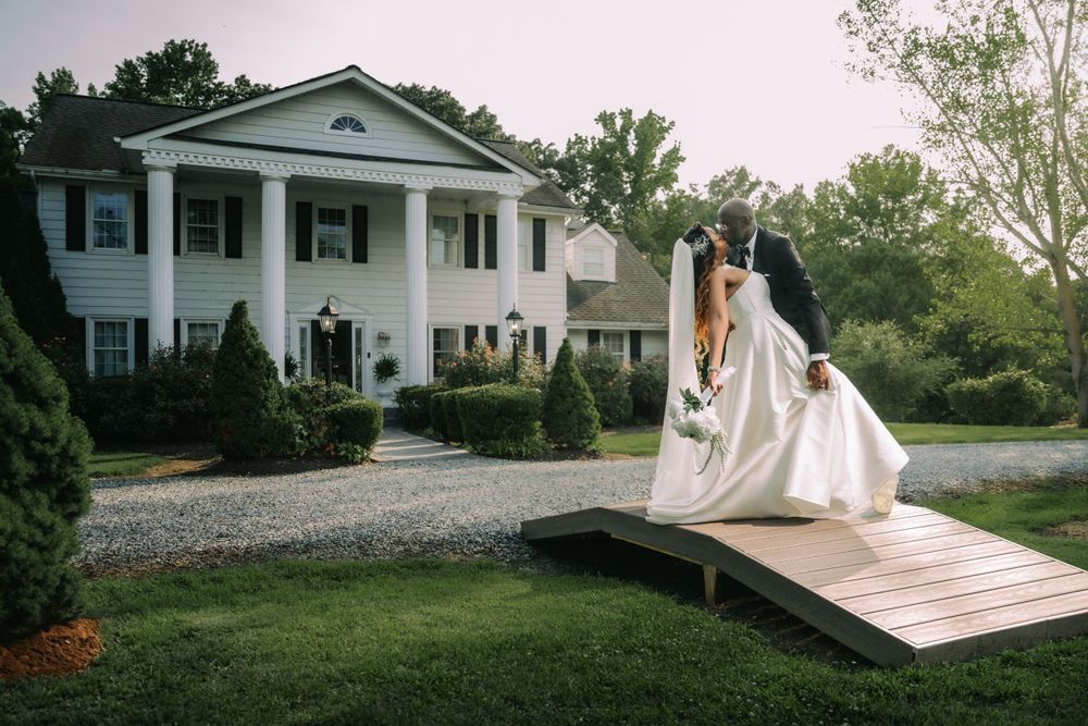 A couple in wedding attire embrace on a wooden bridge in front of a stately white colonial mansion with columns.