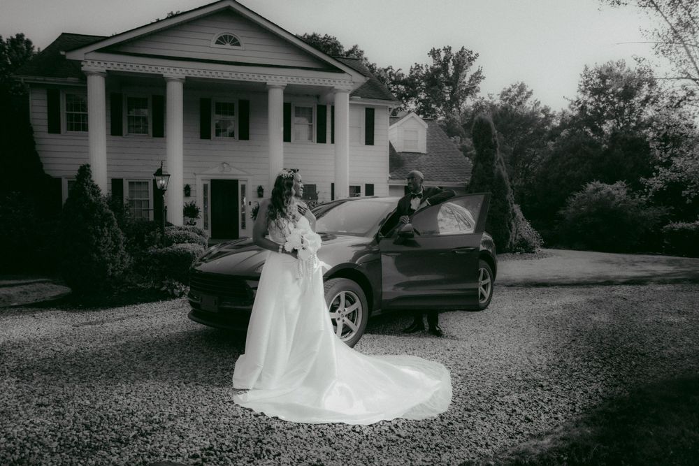 Black and white wedding photo showing someone in a flowing white gown beside a car in front of a grand colonial mansion.