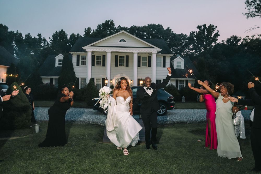 Wedding party celebrates with sparklers outside a stately white mansion with columns during twilight hour.