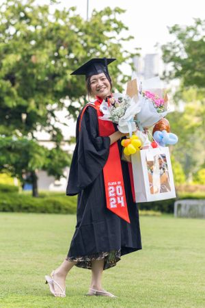 Graduate in black gown and red sash holding bouquets and gifts while standing on grass.