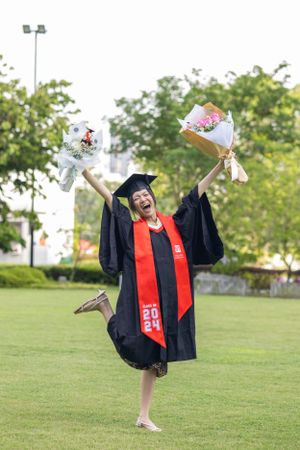 Graduate jumps joyfully holding flower bouquets while wearing academic regalia.