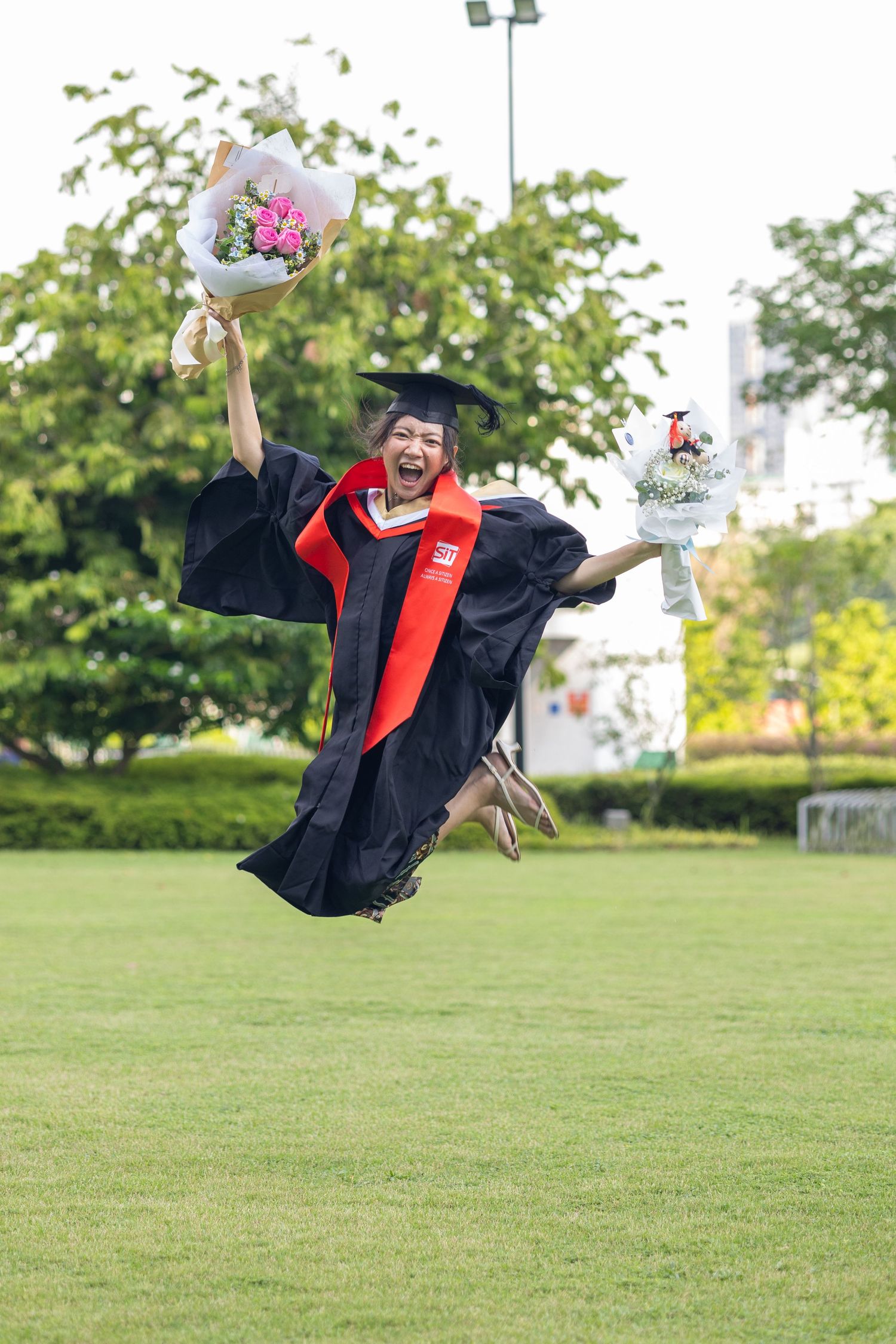Graduate in cap and gown leaping with bouquets on green lawn.