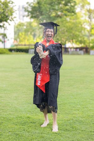 Graduate in academic regalia celebrating with champagne spray on grass.