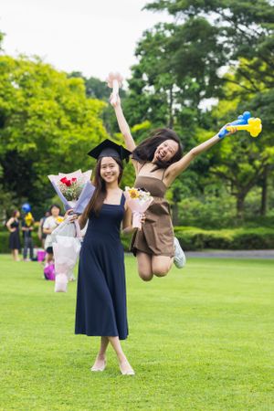 Two graduates celebrating on grass with bouquets and playful poses.