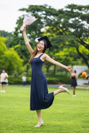 Graduate in navy dress and cap celebrating on grass with raised bouquet and joyful expression.
