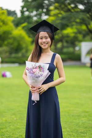 Graduate in navy dress and cap holding pink flower bouquet while standing on green lawn.