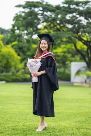 Graduate in traditional black gown and cap holding flower bouquet on grass field.