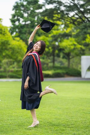 Graduate in black robe raises cap joyfully while striking playful pose on grass.