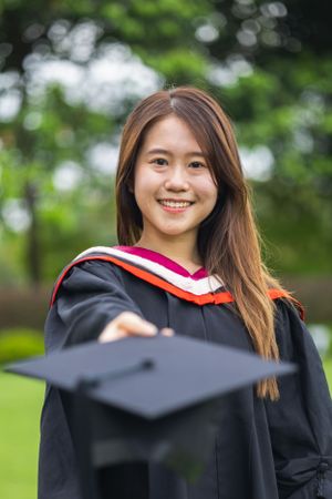 Graduate in black robe tossing graduation cap with natural greenery in background.