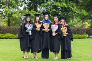 Group of graduates in black robes holding flower bouquets while posing together on lawn.