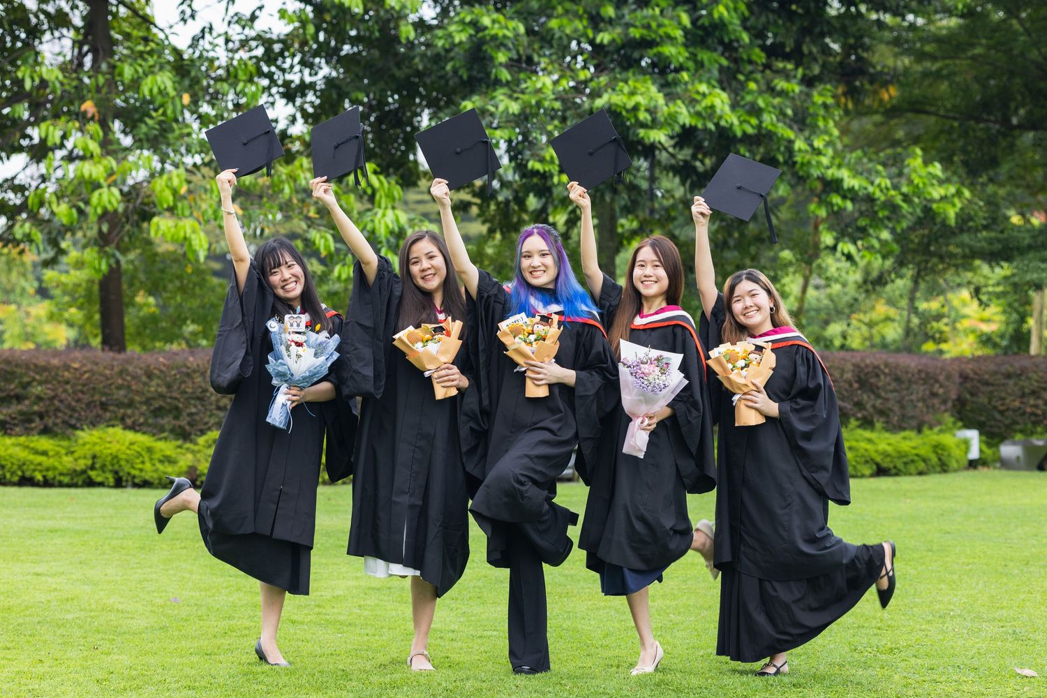 Graduates in black robes jumping with bouquets and caps on green lawn.