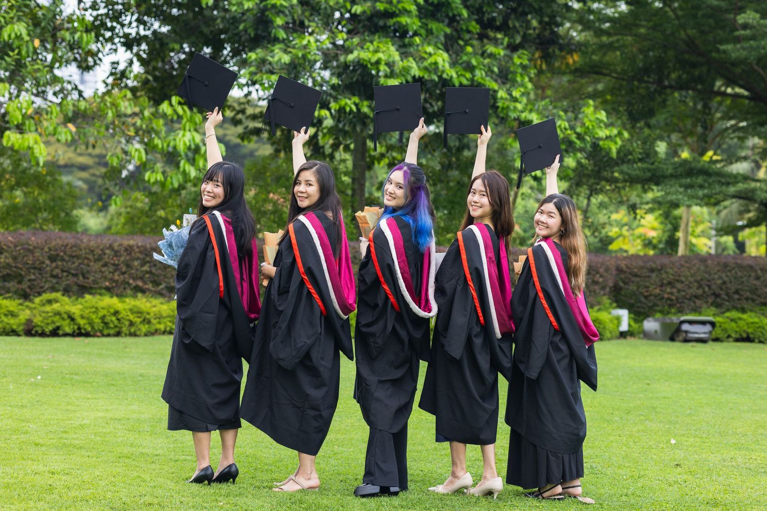 Five graduates in black robes raising caps in celebration on grassy field.