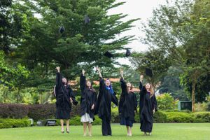 Group of graduates jumping in celebration on campus lawn with trees in background.