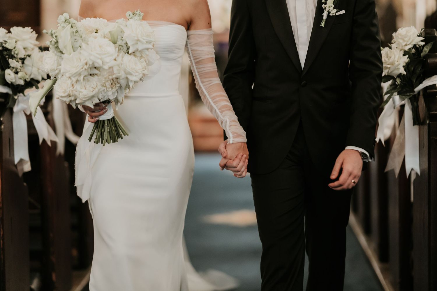 Newlyweds holding hands with white floral bouquet and classic black tuxedo in church aisle.