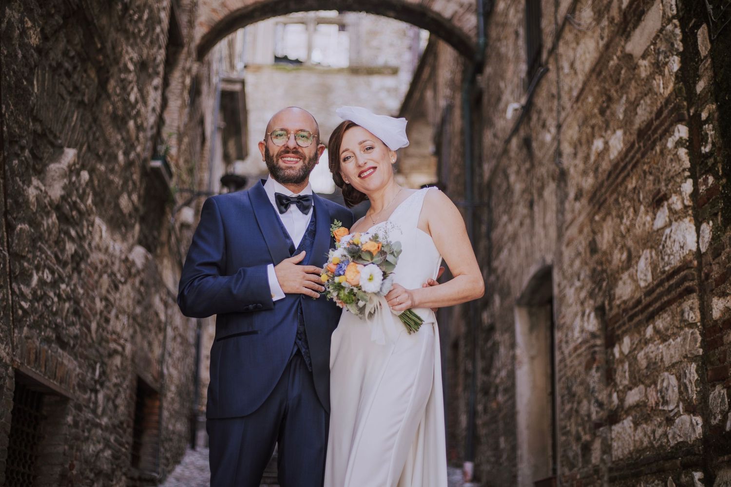 Wedding couple standing in a stone archway alley holding beautiful floral bouquet in classic attire.